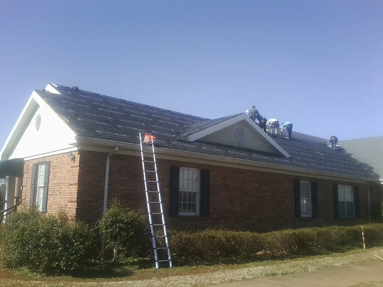 Crew tearing shingles off a brick ranch roof with a ladder set — Metal Roof Repair work by our crew in Rising Fawn