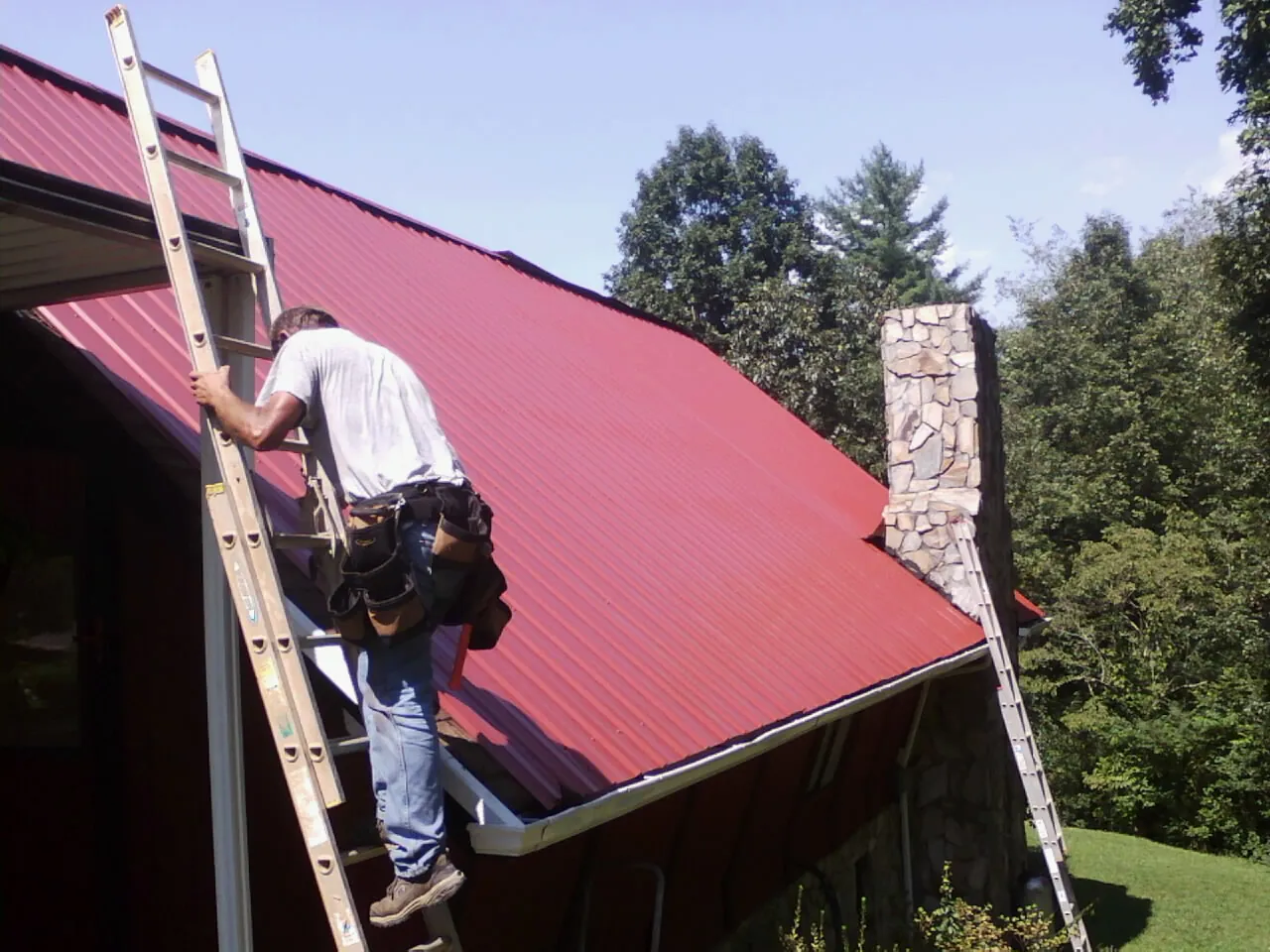 Roofer on a ladder beside a red metal roof and stone chimney — Soffit & Fascia Repair by Roofing Brothers in Rising Fawn